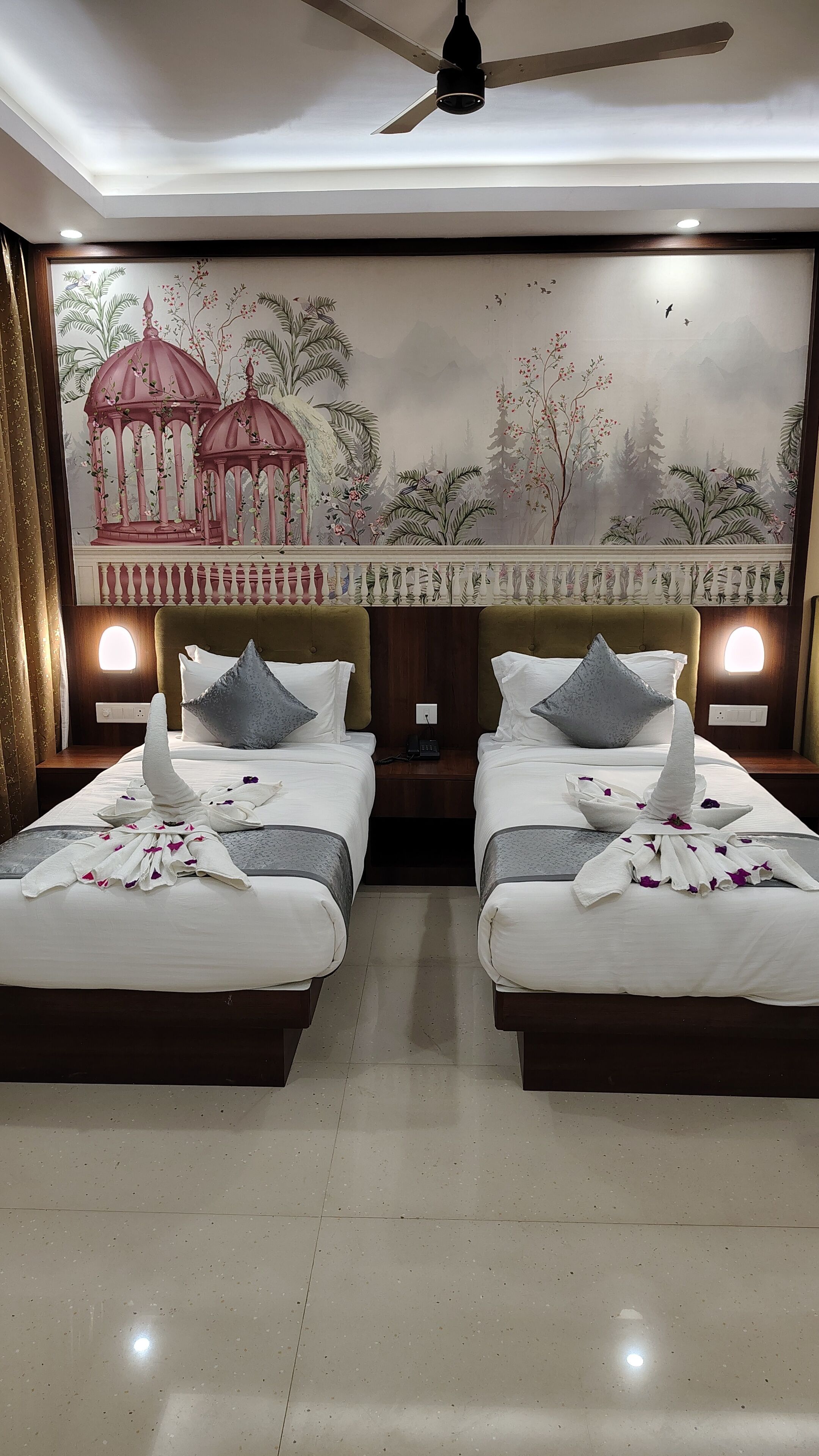 Quarto de hotel com duas camas, um ventilador de teto, um mural na parede e luminárias de cabeceira.