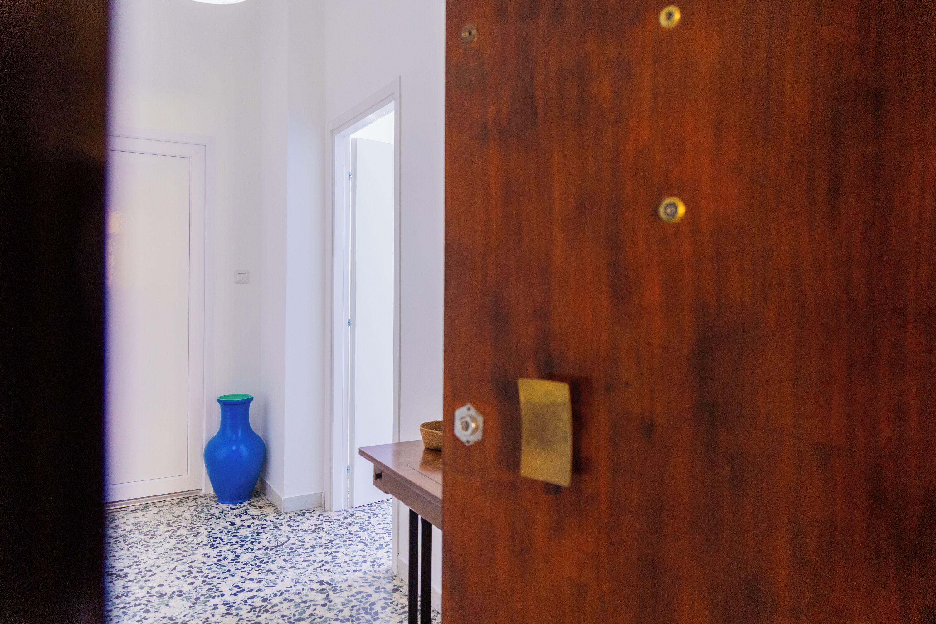 Un pasillo con una puerta de madera, un jarrón azul y piso de baldosas.