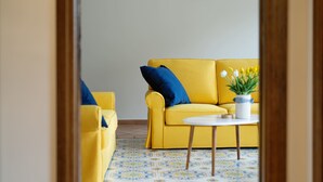 Un divano giallo con cuscini blu, un tavolo bianco e una pianta in vaso in una stanza con pavimento piastrellato.