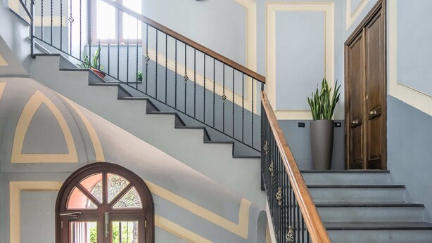 Un'imponente scalinata con ringhiera in legno e balaustre nere, che conduce a una porta con arco decorativo.