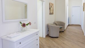 Un intérieur moderne avec une coiffeuse blanche, un miroir, un vase de fleurs, un fauteuil beige et un tableau au mur.