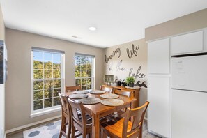 Un coin repas avec une table en bois dressée pour six personnes, un réfrigérateur blanc et un mur orné d’une citation inspirante.