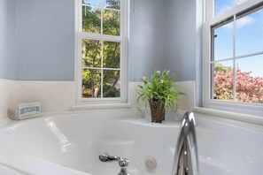 Une salle de bain avec une baignoire blanche, deux fenêtres et une plante en pot.