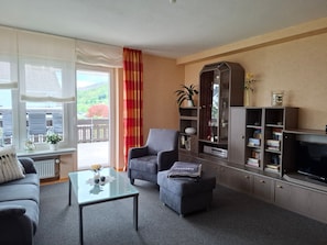 A living room with a sofa, armchair, ottoman, glass coffee table, and a TV stand with bookshelves. - Holiday Apartment 'Waltraud, 105 sqm' with Panoramic View, Private Terrace & Wi-Fi (Brilon)