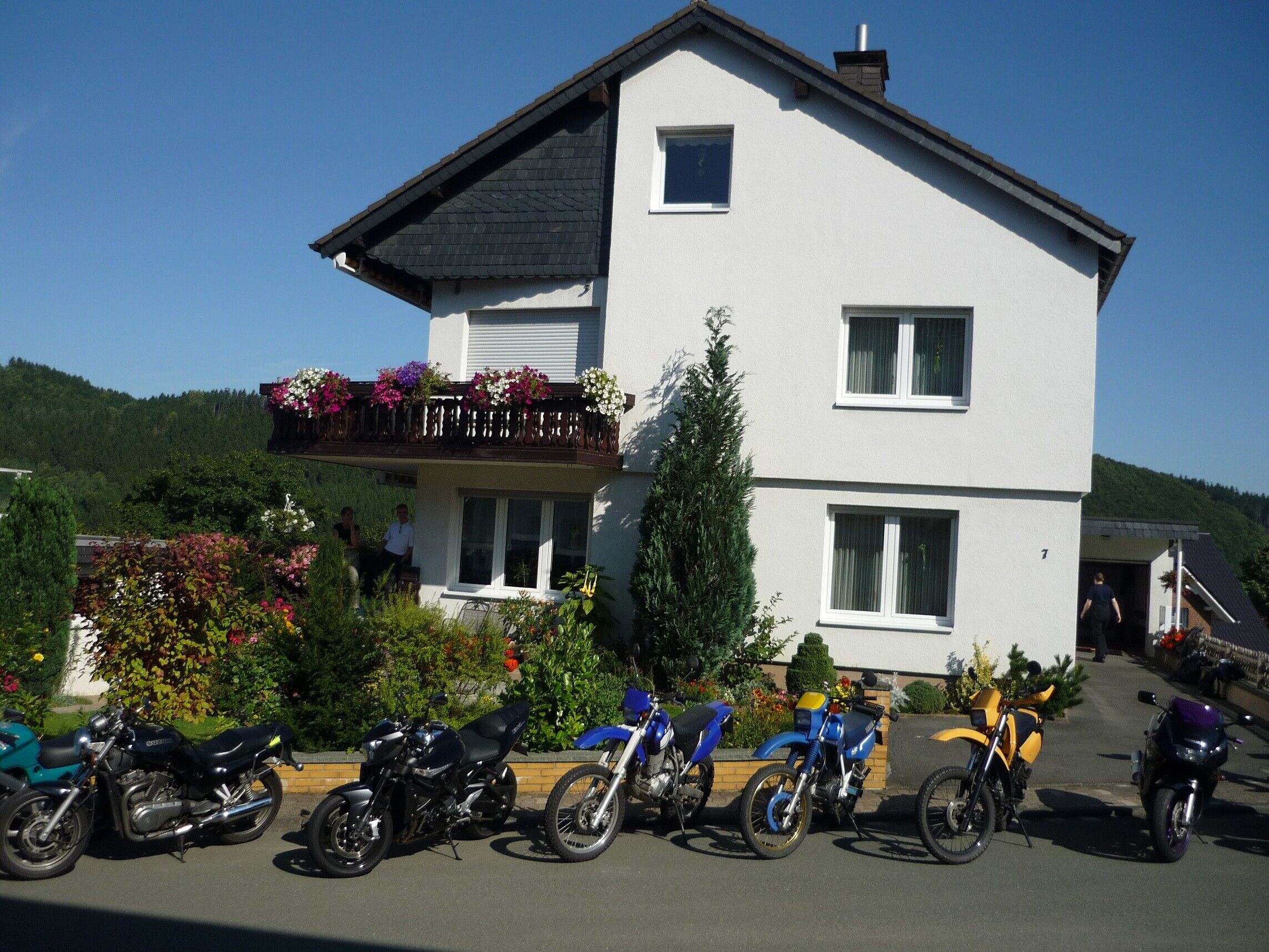 Un bâtiment à deux étages doté d’un balcon, un espace de stationnement où sont garées des motos, et un paysage environnant.