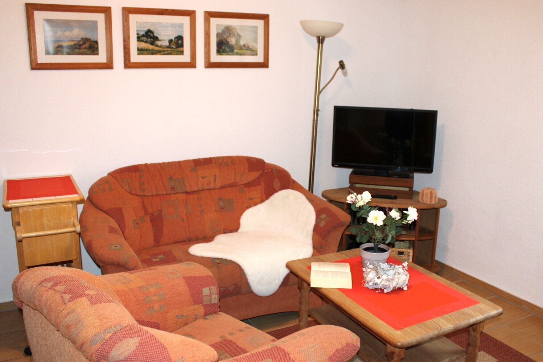 Sala de estar com um sofá, uma mesinha com um vaso de flores, uma televisão e quadros nas paredes.