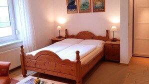 Ein hölzernes Bett mit zwei Nachttischen, ein Fenster mit Vorhängen und drei Reiseplakate an der Wand.
