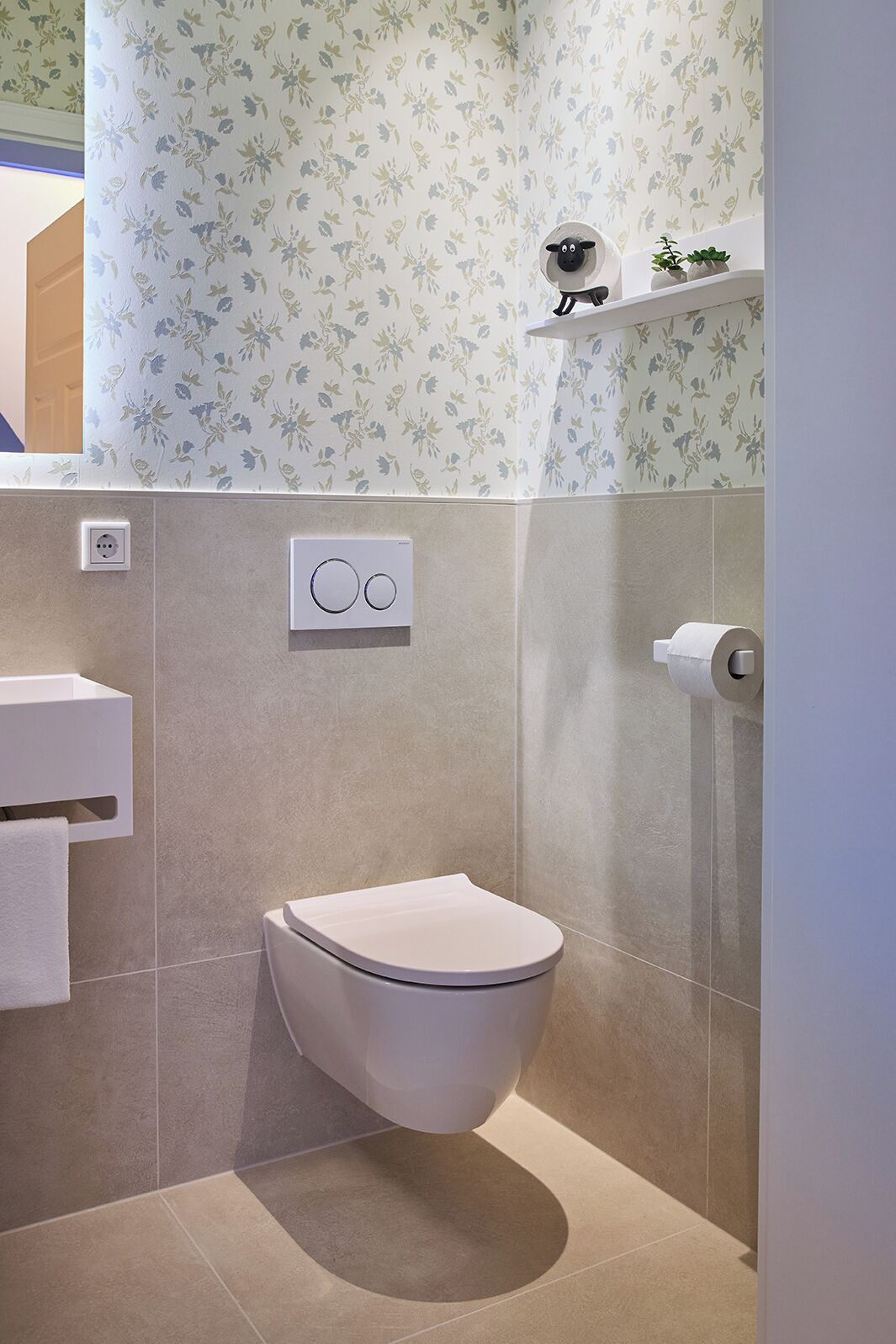 Casa de banho com sanita, porta-toalhas, prateleira com plantas e botão de descarga na parede.