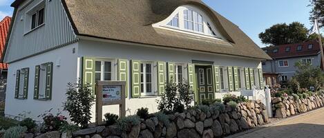 Ein traditionelles Gebäude mit einem strohgedeckten Dach, grünen Fensterläden und einer Mauer im Vordergrund.