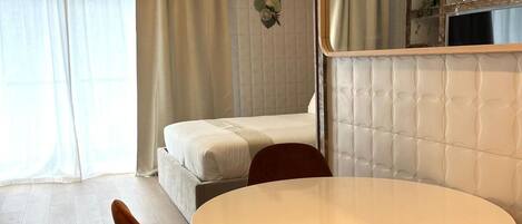 Quarto de hotel moderno com uma mesa de jantar redonda, duas cadeiras laranja, uma cama, um espelho grande e um detalhe decorativo na parede.