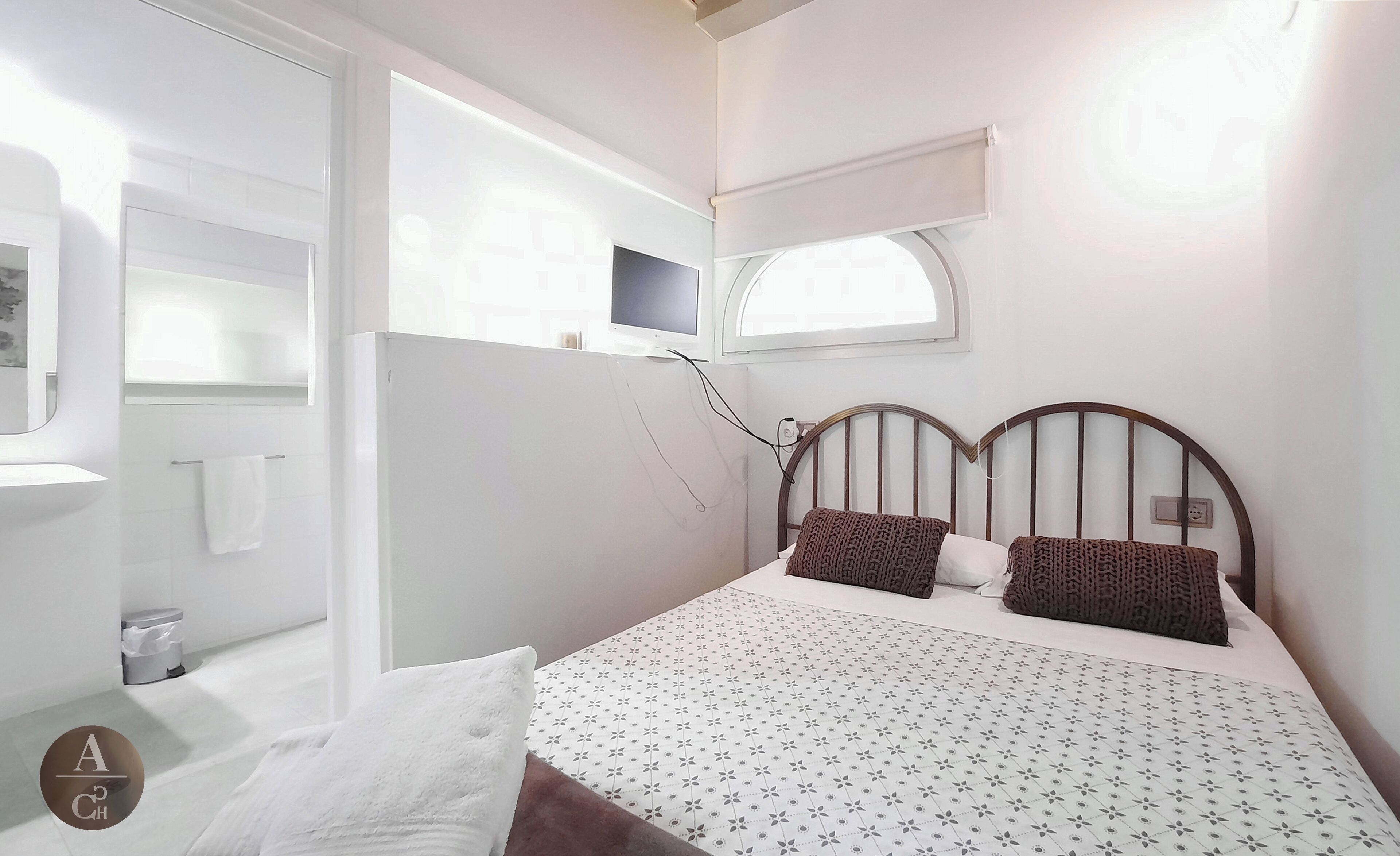 Un dormitorio con cama, dos almohadas, televisor, ventana y baño a la vista.