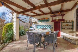 Um pátio externo com mesa e cadeiras, teto de madeira e área de cozinha com prateleiras e uma porta vermelha.