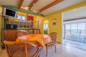 Uma área de refeições com mesa e cadeiras, uma televisão fixada numa viga de madeira, e uma janela com vista.