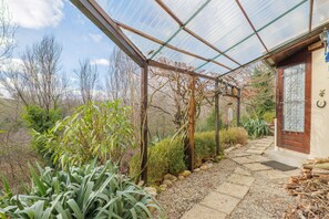 Una estructura invernadero con armazón de madera y paneles de vidrio, rodeada de diversas plantas y un sendero de piedra que lleva a una puerta.