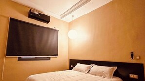 A hotel room with a bed, a flat-screen TV mounted on the wall, a bedside table, and a wall-mounted light fixture. - Relax family Majestic (Ouarzazate)