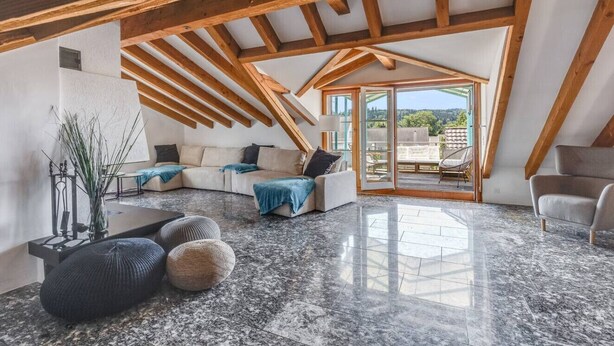 Een ruime loft met een hoge plafond, zichtbare houten balken, een groot raam en een gezellige zithoek met een bank en poefs.