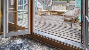 Een ruim balkon met een houten terras, een glazen deur en een overdekte zithoek met een stoel en een tafel.