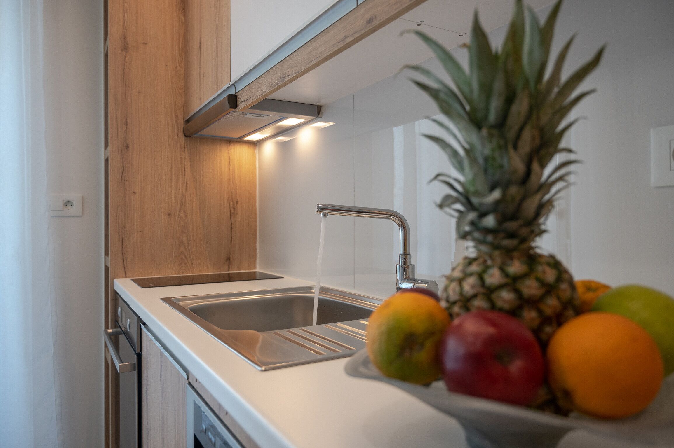 A kitchen with a stainless steel sink, wooden cabinetry, and a bowl of fruit on the counter.