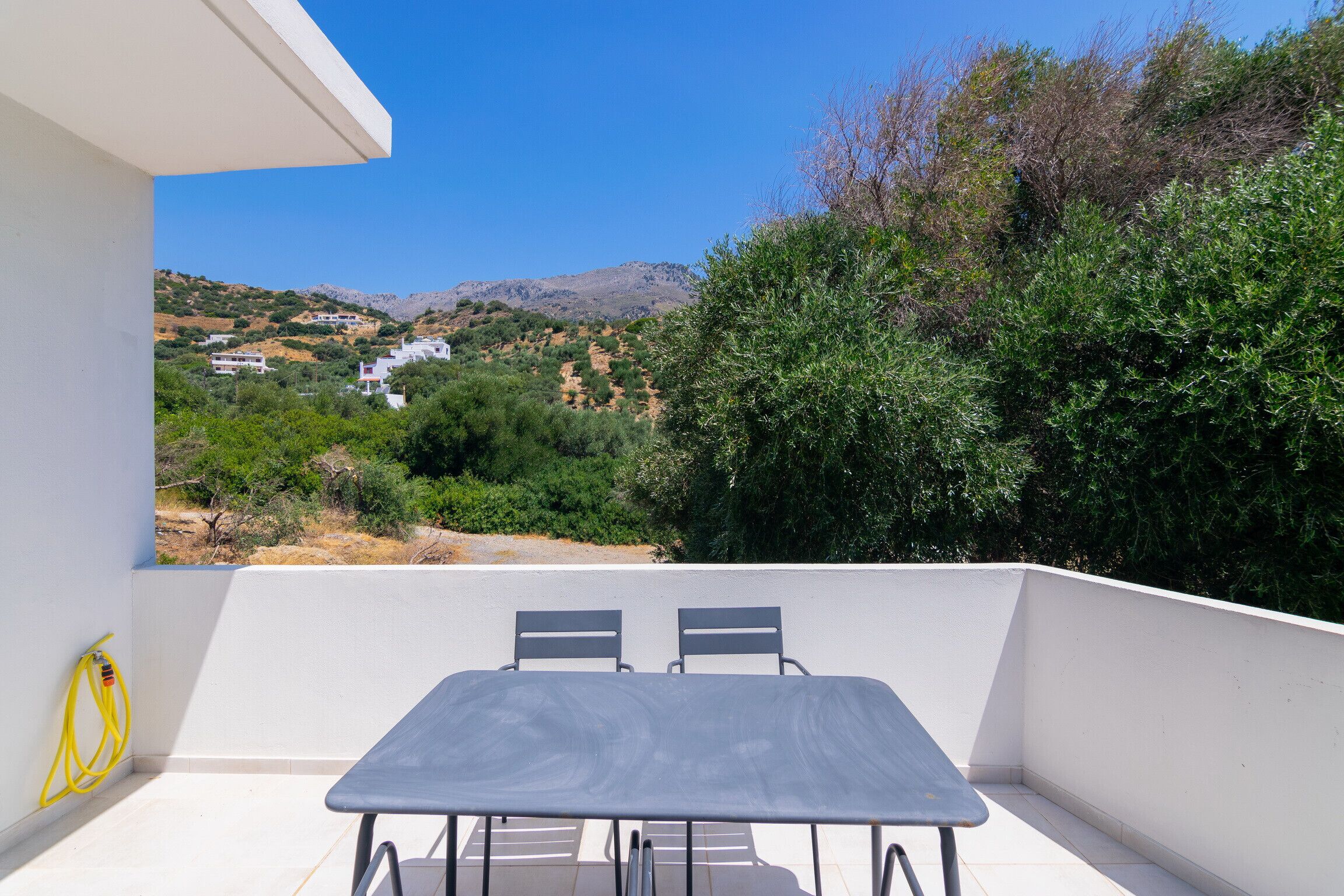 A balcony with a table and chairs, overlooking a hillside with buildings and greenery.