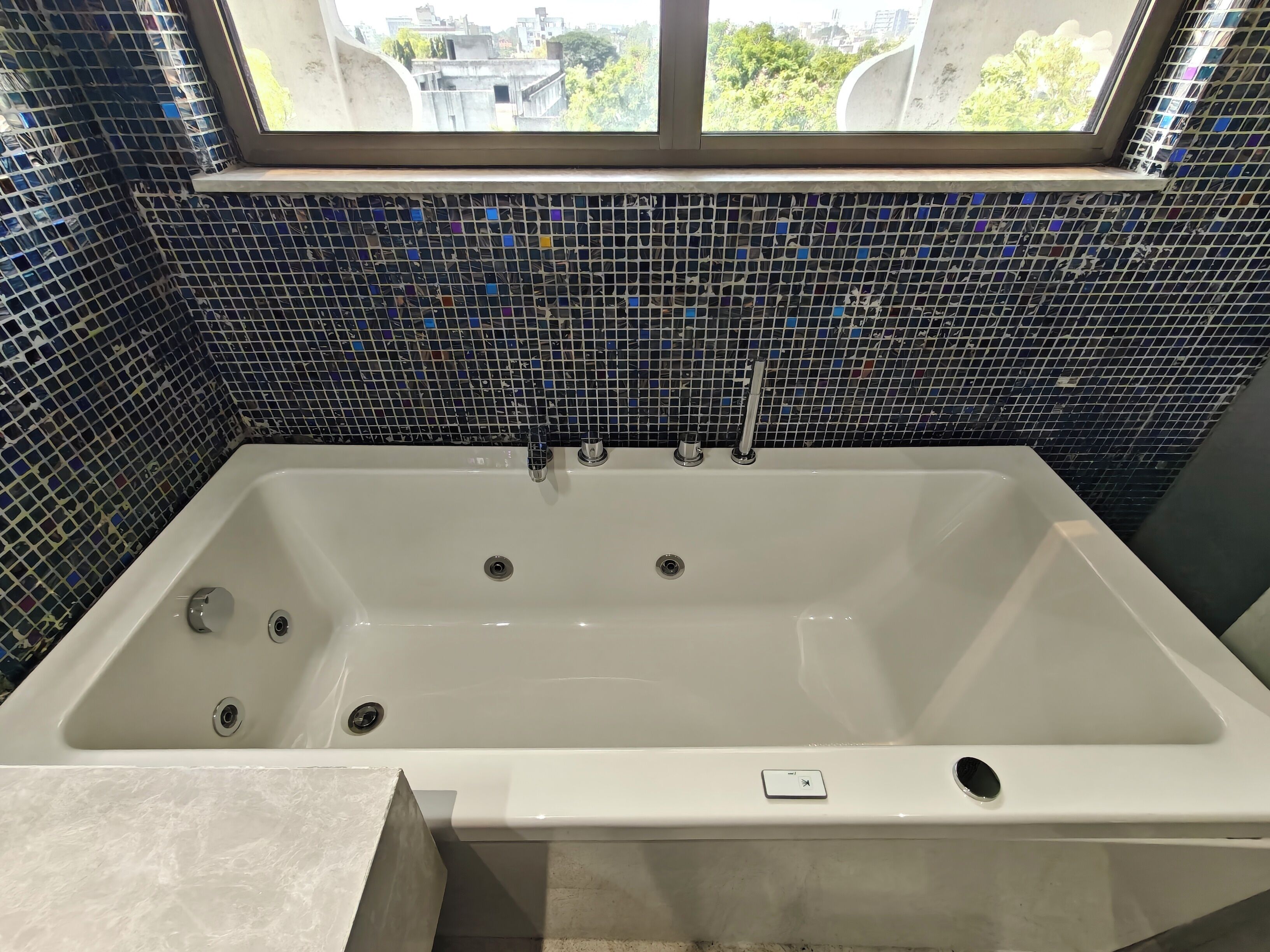 A modern bathroom with a large bathtub, a window above it, and a mosaic tile wall.