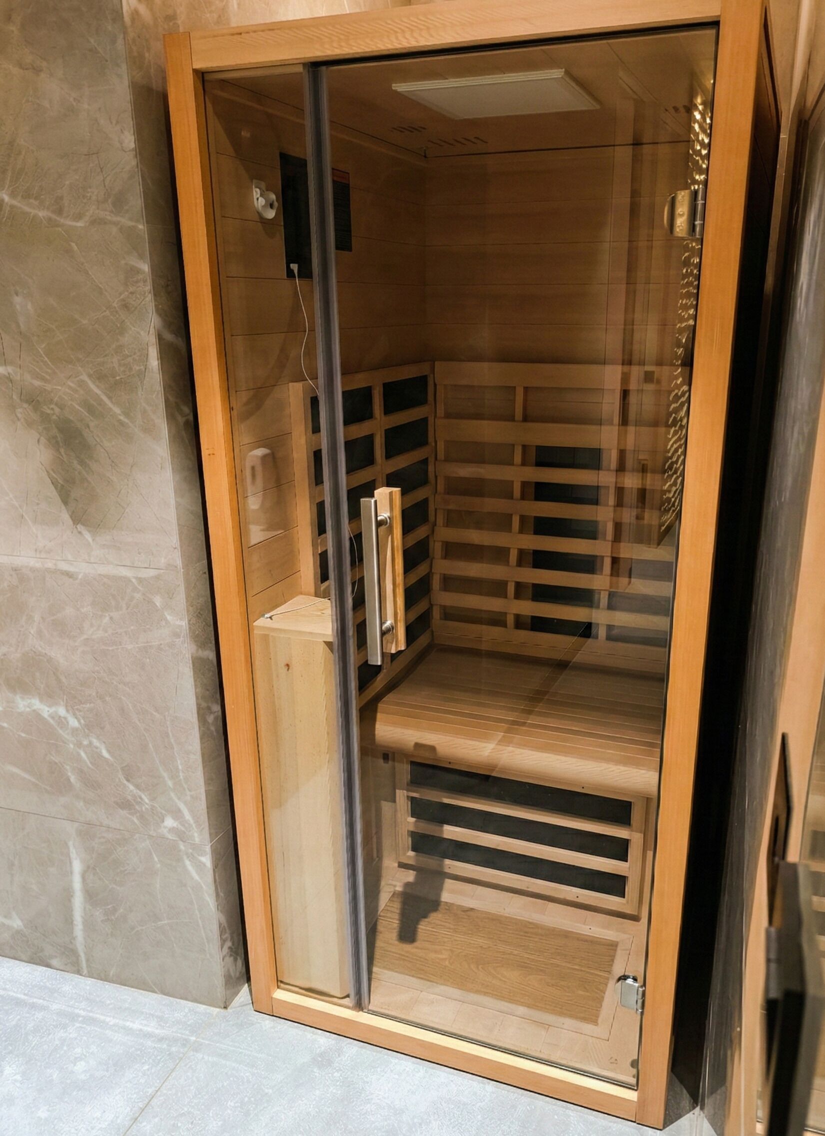 Uma sauna com interior em madeira, porta de vidro e puxador de metal.
