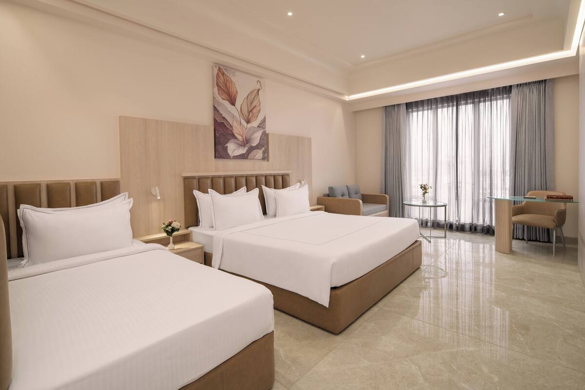 Habitación de hotel con dos camas, un escritorio, una silla, una mesita pequeña y un ventanal con cortinas.
