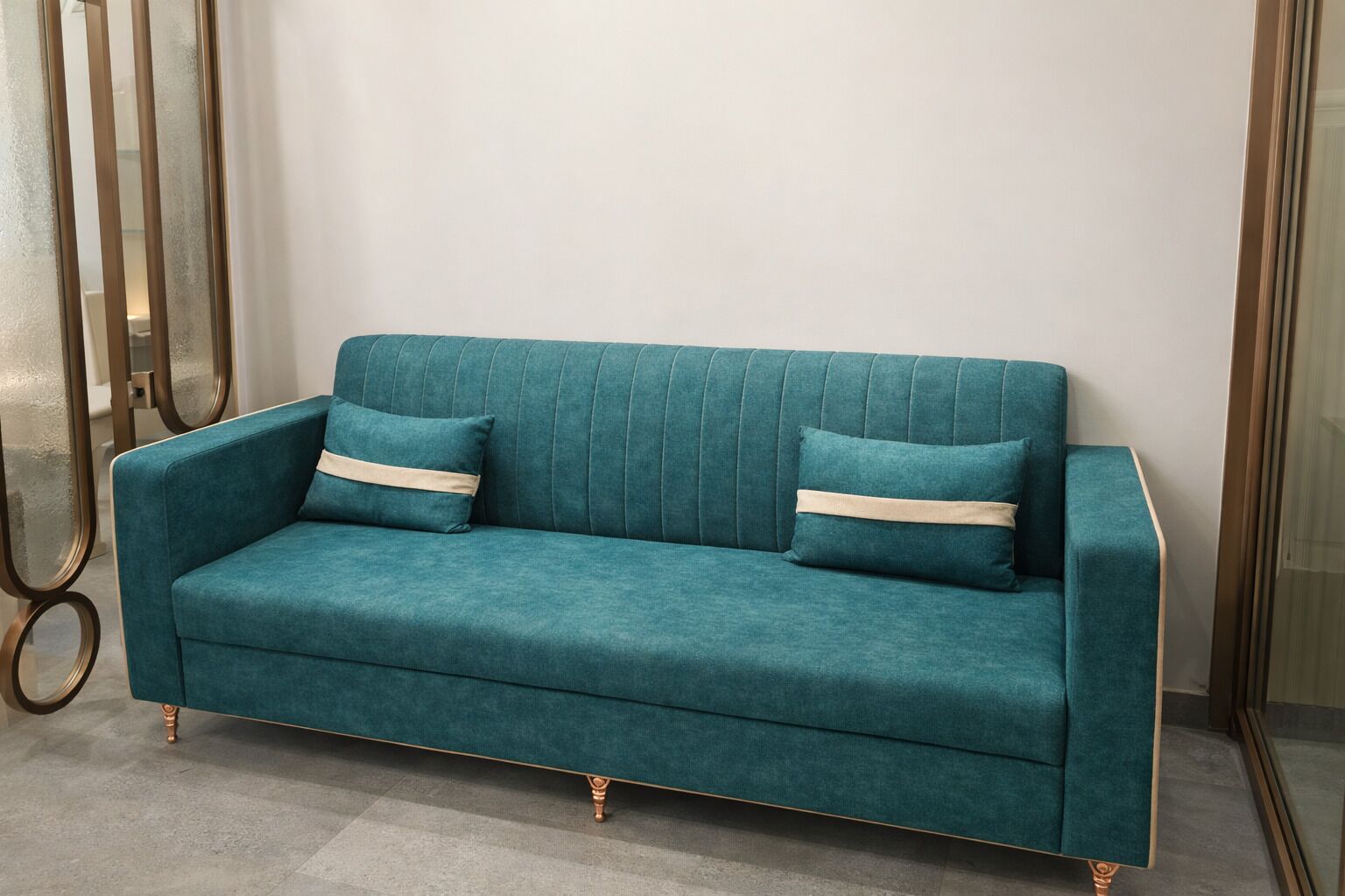 Un canapé de couleur turquoise avec des pieds dorés et deux coussins beige, dans une pièce dotée d’une cloison en verre et d’une structure métallique sur le côté gauche.