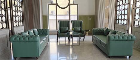 Un vestíbulo amplio con dos sofás de cuero verde, una mesa de centro redonda de cristal y un sillón verde. Un gran ventanal proporciona luz natural.