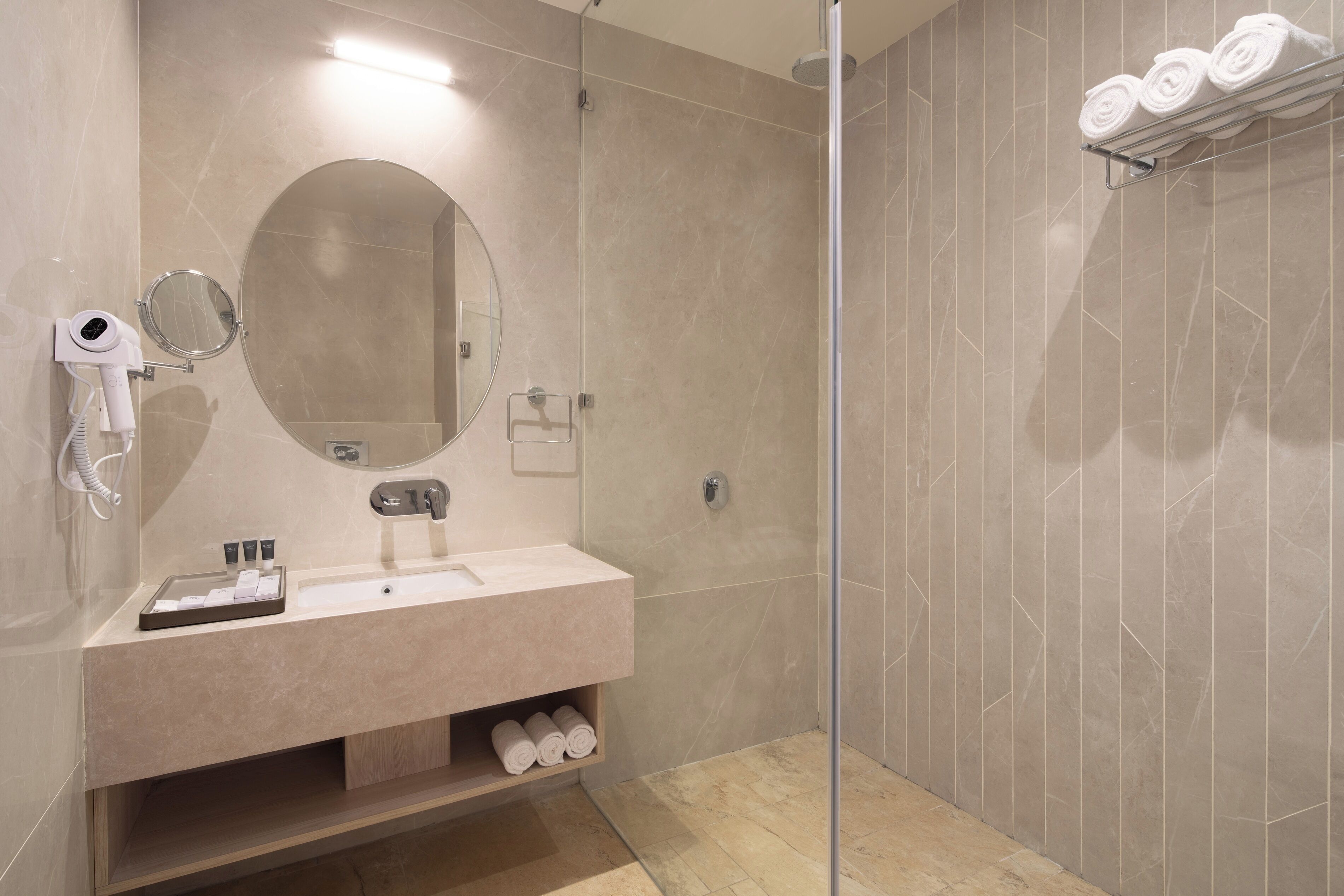 A modern bathroom with a glass-enclosed shower, a round mirror, a sink with toiletries, and a towel rack.