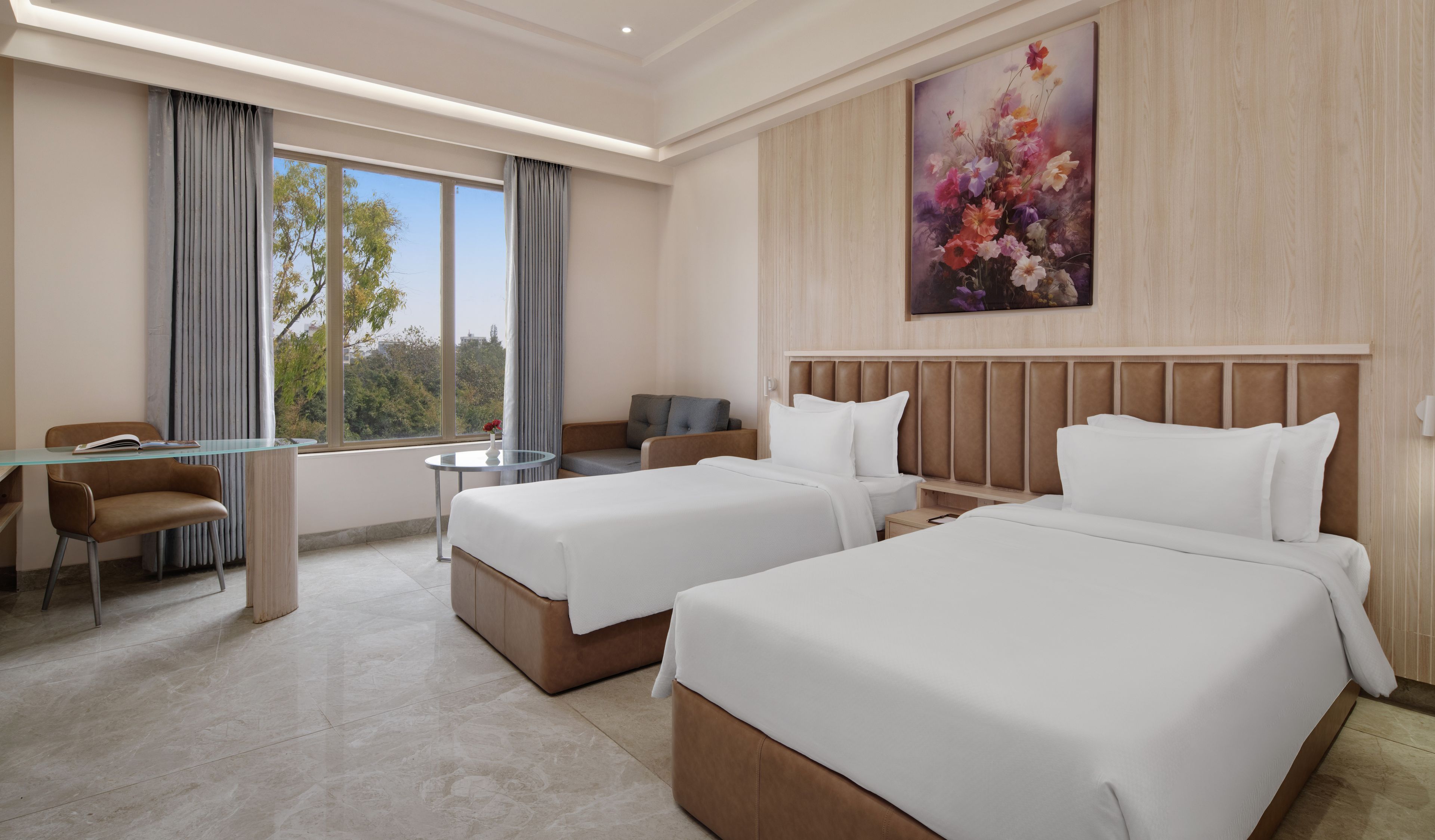 A hotel room with two beds, a desk, a chair, and a view of trees through large windows.