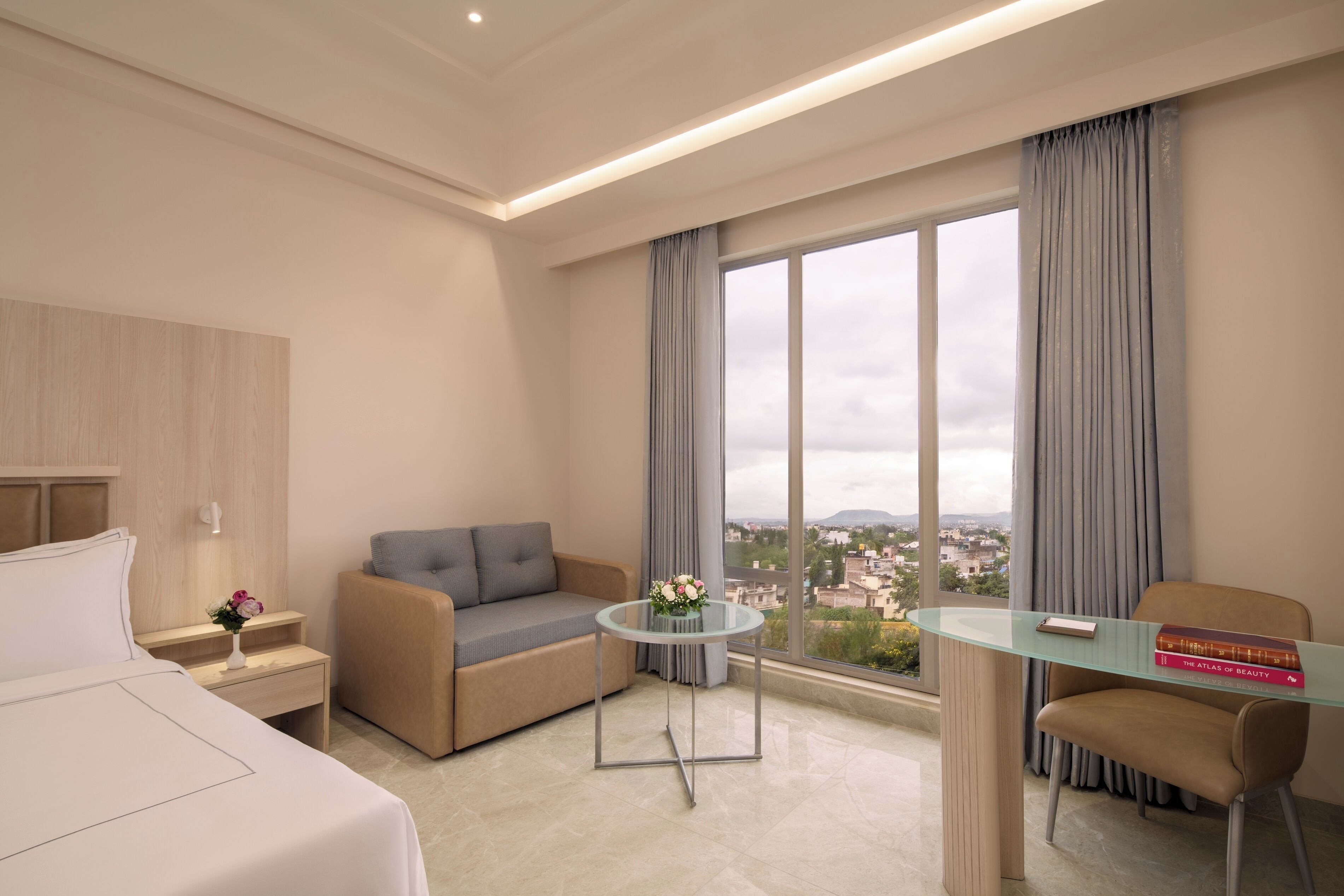 Un hotel moderno con cama, sofá, una mesita con un florero, una silla y vistas de la ciudad a través de amplias ventanas.