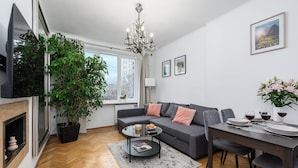 A modern living room with a grey sofa, a coffee table, a dining table set for two, and a large potted plant. - Lumina apart near Royal Lazienki Park (Warsaw)