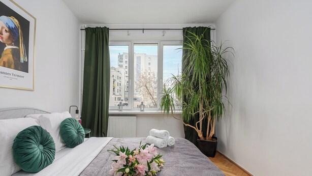 A neatly arranged bedroom with a bed, pillows, a potted plant, and a window with curtains. - Lumina apart near Royal Lazienki Park (Warsaw)