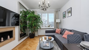 A modern living room with a grey sofa, a round coffee table, a TV, and a large window with a view of buildings. - Lumina apart near Royal Lazienki Park (Warsaw)