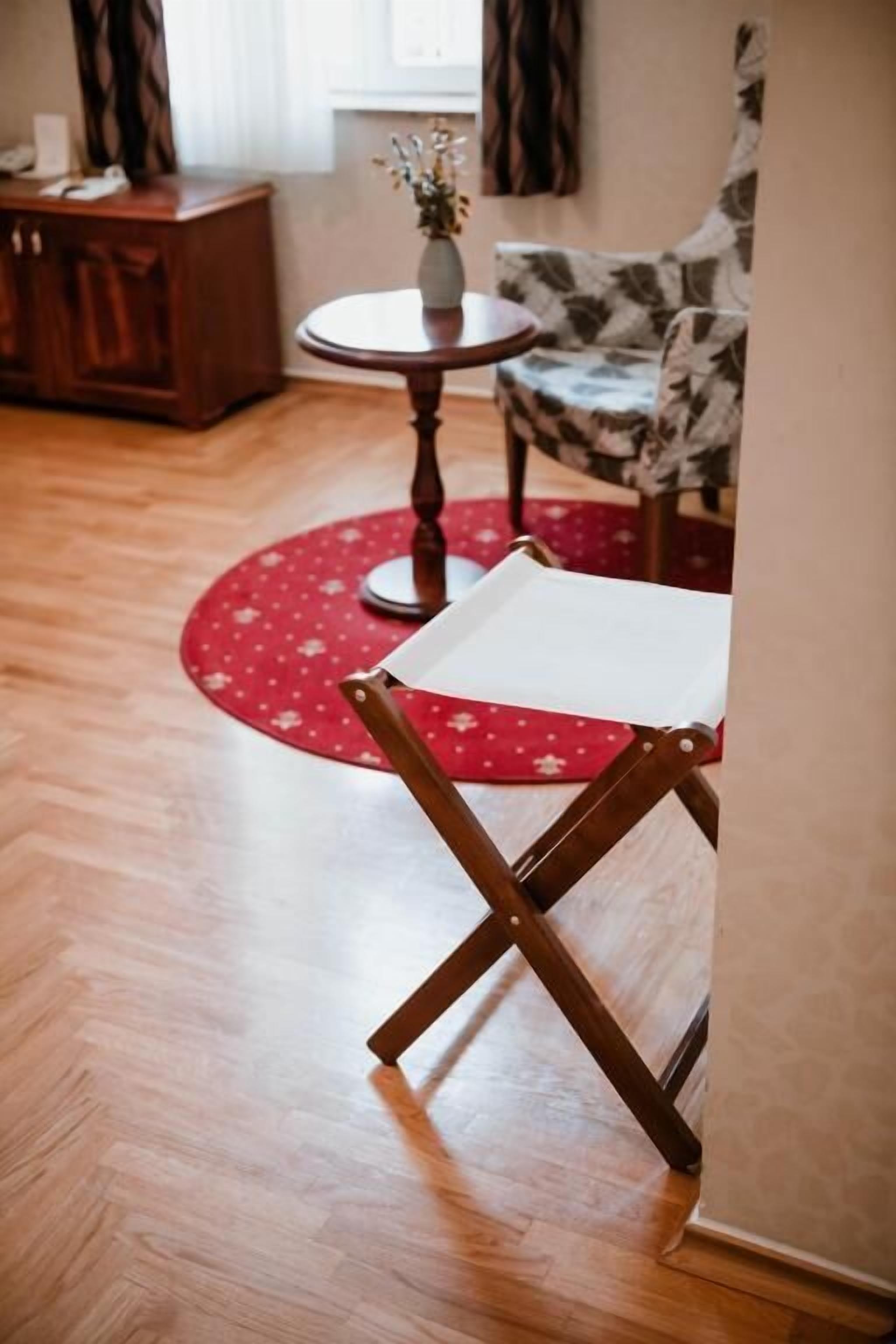 A room with a wooden floor, a red patterned rug, a small round table with a vase, and a floral-patterned chair.