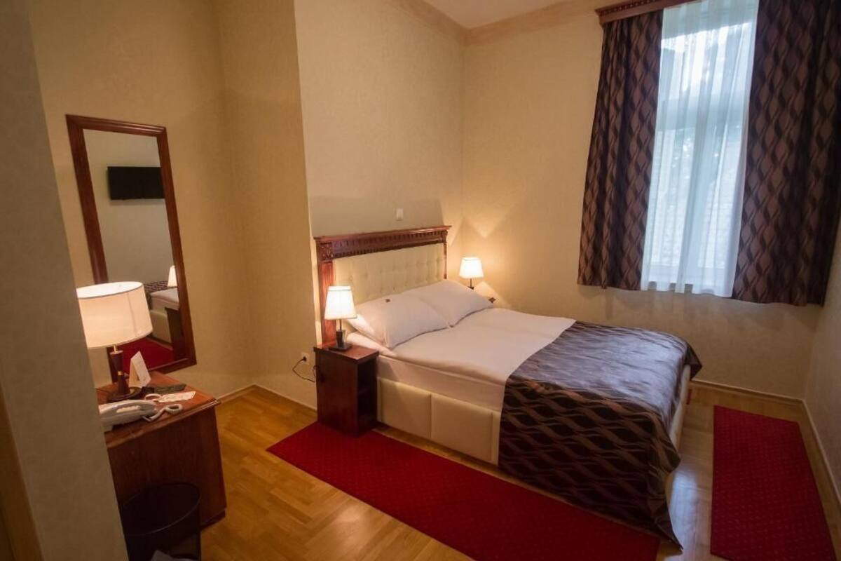 Habitación de hotel con cama, lámparas de noche, un espejo, un escritorio con teléfono y una alfombra roja.