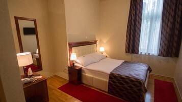 Een hotelkamer met een bed, bedlampen, een spiegel, een bureau met telefoon en een rood vloerkleed.