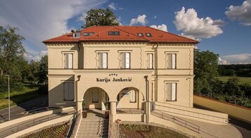 Edificio de dos pisos con techo rojo, llamado "Kurija Janković", que presenta una entrada central con arcos y un pequeño jardín delantero.