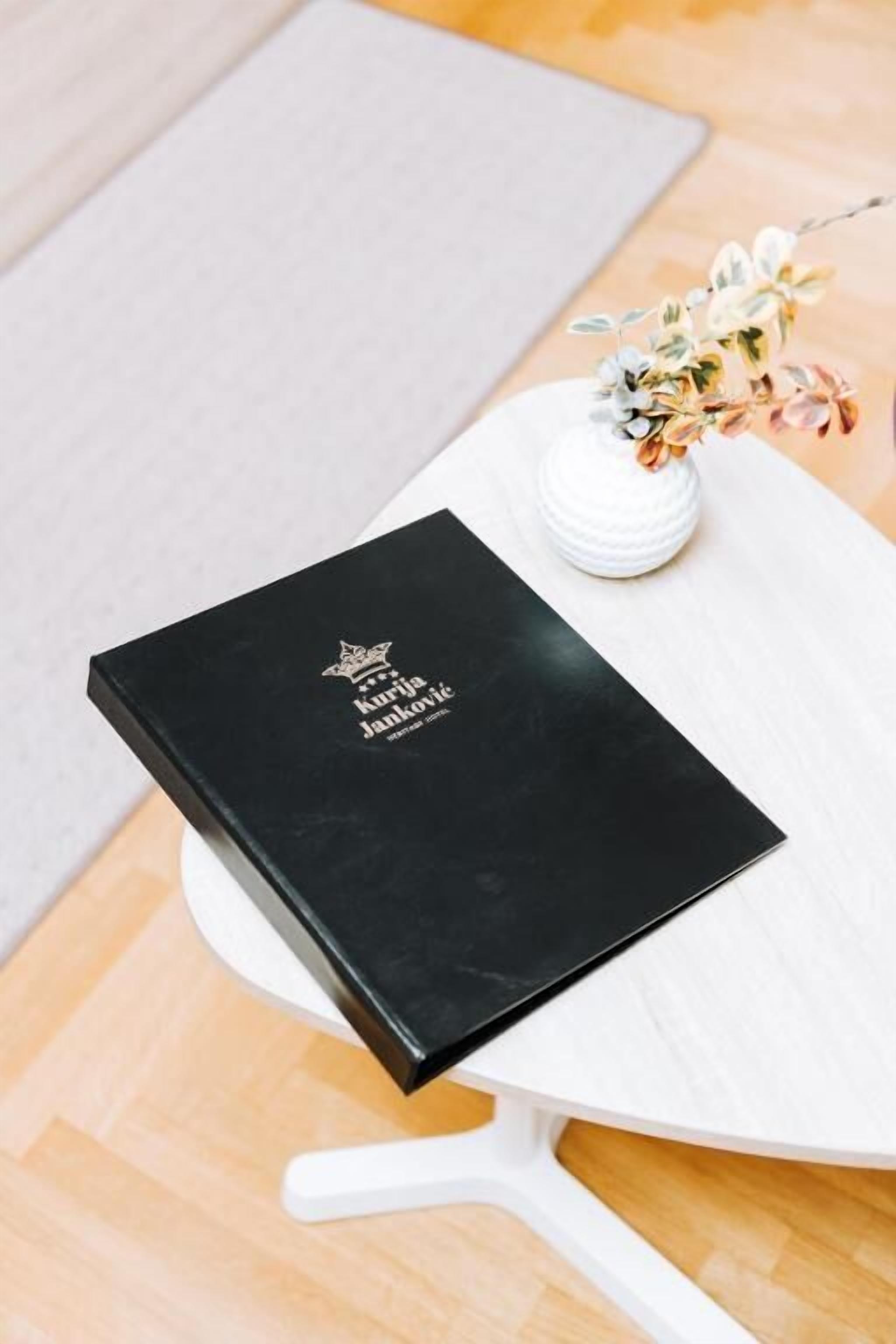 Een zwart boek met de titel 'Kurija Janković' op een witte tafel, met een witte vaas met bloemen.