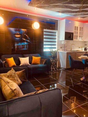 A modern living room with a black sofa, a dining area with a blue chair, and a kitchen with white cabinets. - AzureGold Apartment (Senglea)