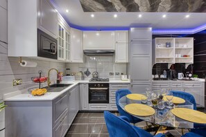 A modern kitchen with white cabinetry, a built-in oven, and a dining area with blue chairs and a glass table. - AzureGold Apartment (Senglea)