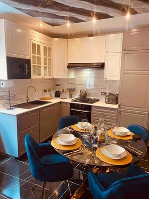 A modern kitchen with white cabinets, a gas stove, and a dining area with blue chairs and gold placemats. - AzureGold Apartment (Senglea)