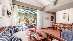 A spacious living area with a sofa, chairs, a glass coffee table, and a dining table with chairs. There is a large window with curtains, a view of trees, and a ceiling with exposed beams.