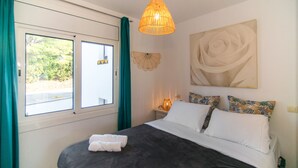 A bedroom with a large bed, a window with curtains, a wall-mounted lamp, and a floral painting. - WELCS APARTMENT 295 EMP with community pool (Rosas / Roses)