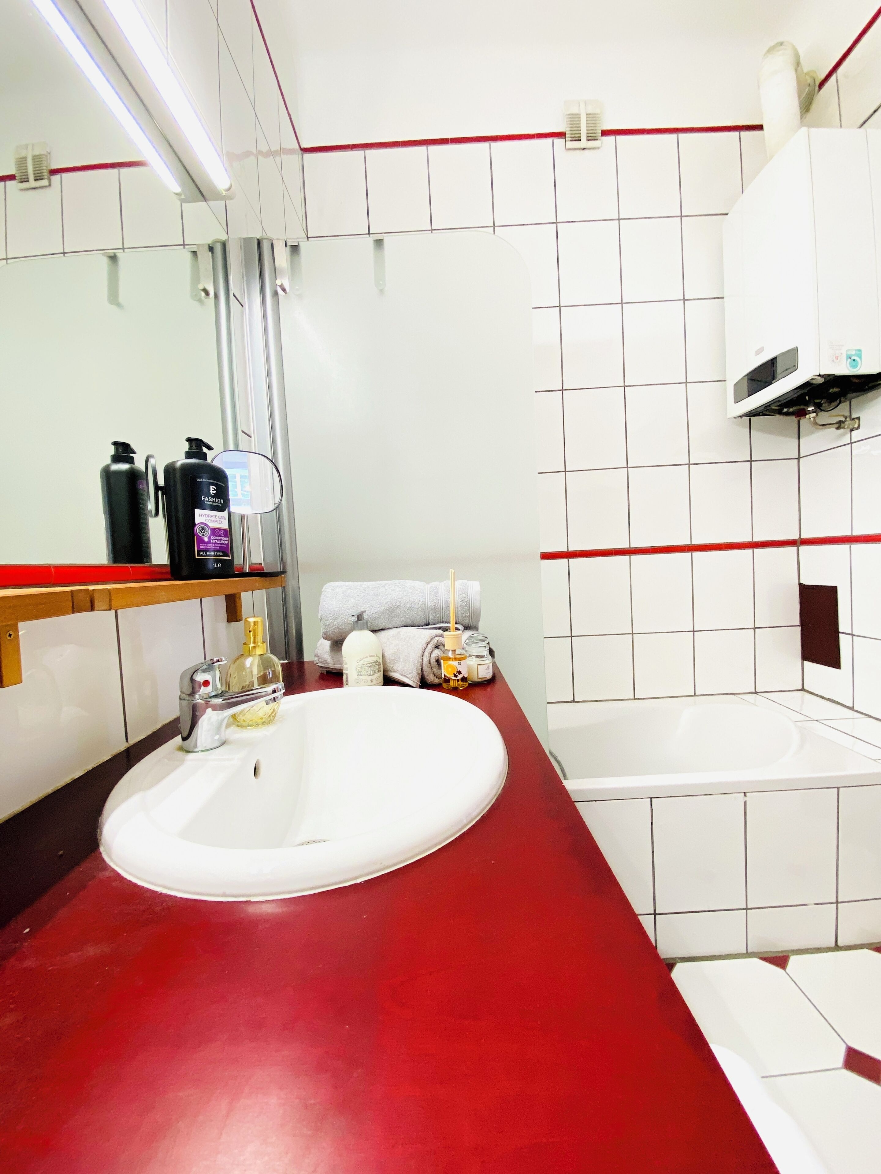 Un baño con lavamanos blanco, encimera roja y un espejo encima. Hay artículos de aseo en la encimera y un perchero con toallas.
