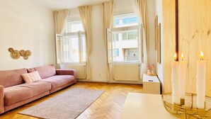A living room with a herringbone wooden floor, a pink sofa, a white side table with a candle holder, and large windows with curtains. - Cozy 2BR Apartment Near Westbahnhof (Vienna)
