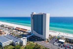 A high-rise hotel with a beach view, parking area, and a pier in the background. - Fall Deals + Snowbird Rates | Gulf Front Beach Escape (Panama City Beach)