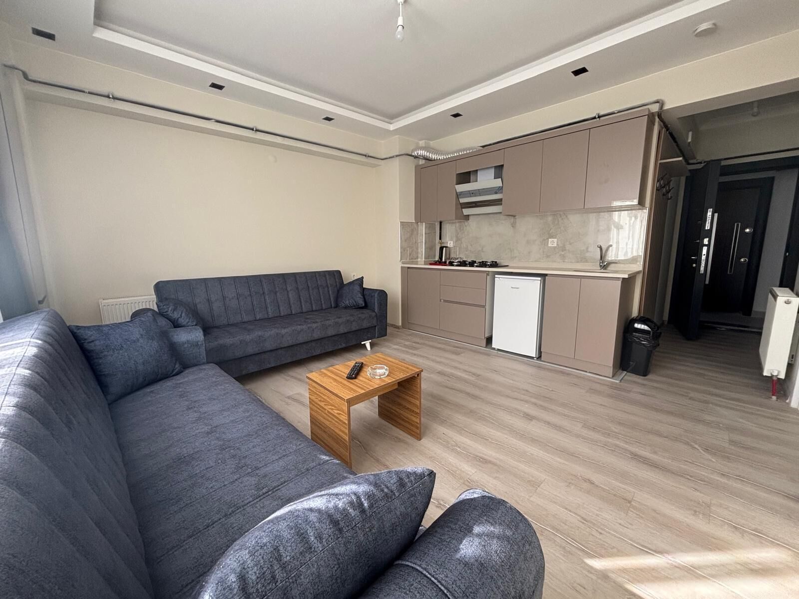 Un salon moderne avec un canapé d’angle gris, une table basse en bois et un coin cuisine doté d’un réfrigérateur blanc et de meubles de rangement de couleur beige.