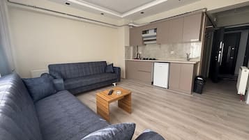 Un salon moderne avec un canapé d’angle gris, une table basse en bois et un coin cuisine doté d’un réfrigérateur blanc et de meubles de rangement de couleur beige.