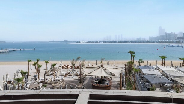 Vista de la playa con zona de bar, palmeras y un horizonte urbano a lo lejos.